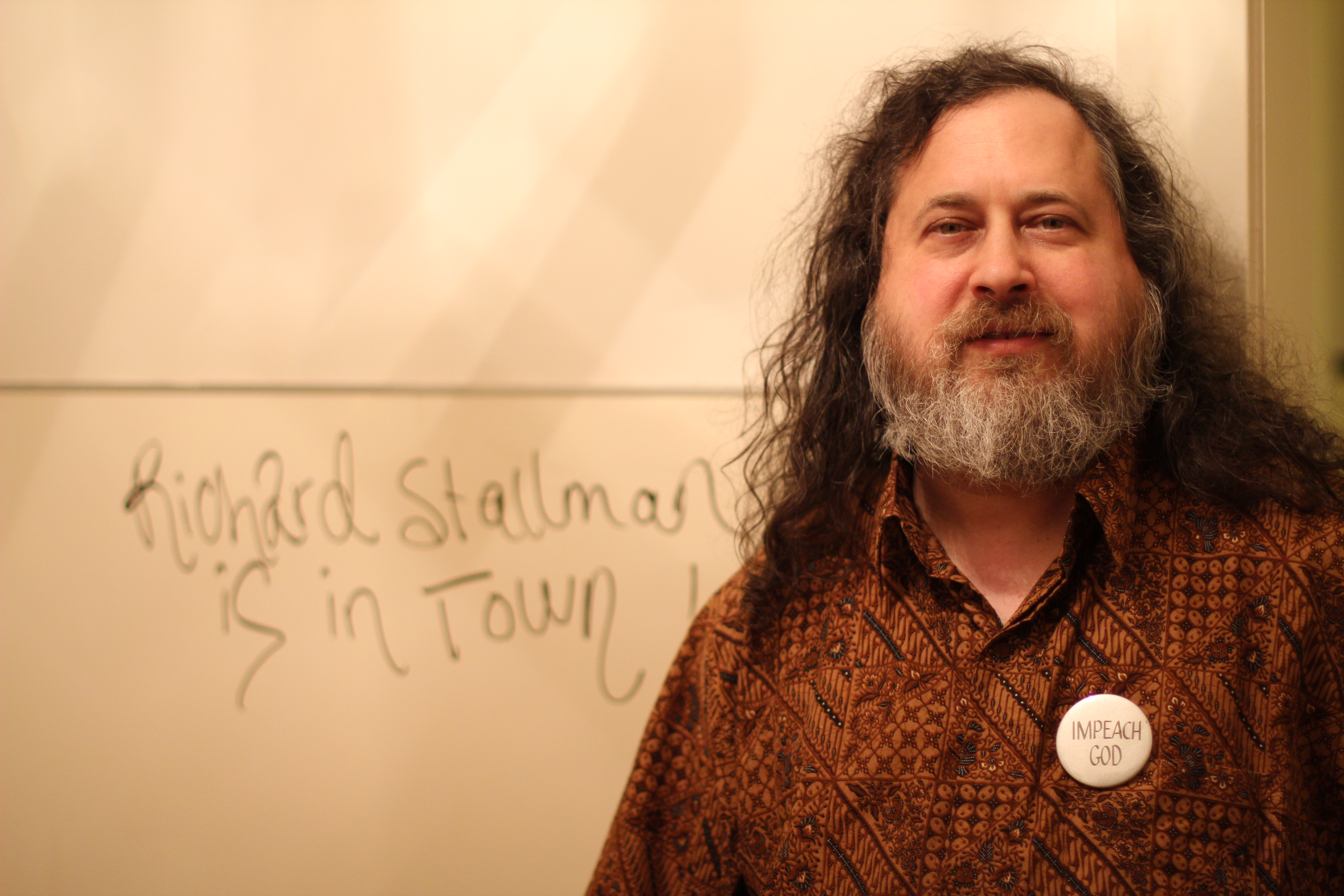 Foto do Richard Stallman, um homem estrutura média, barbudo e cabelos grandes e com um fisico corporal forte. Utilizando uma camisa de botão na cor marrom. Ele está em pé em uma sala de reuniões e ao fundo uma losa escrito - Richard Stallman is in town! - nessas palavras no idioma inglês.